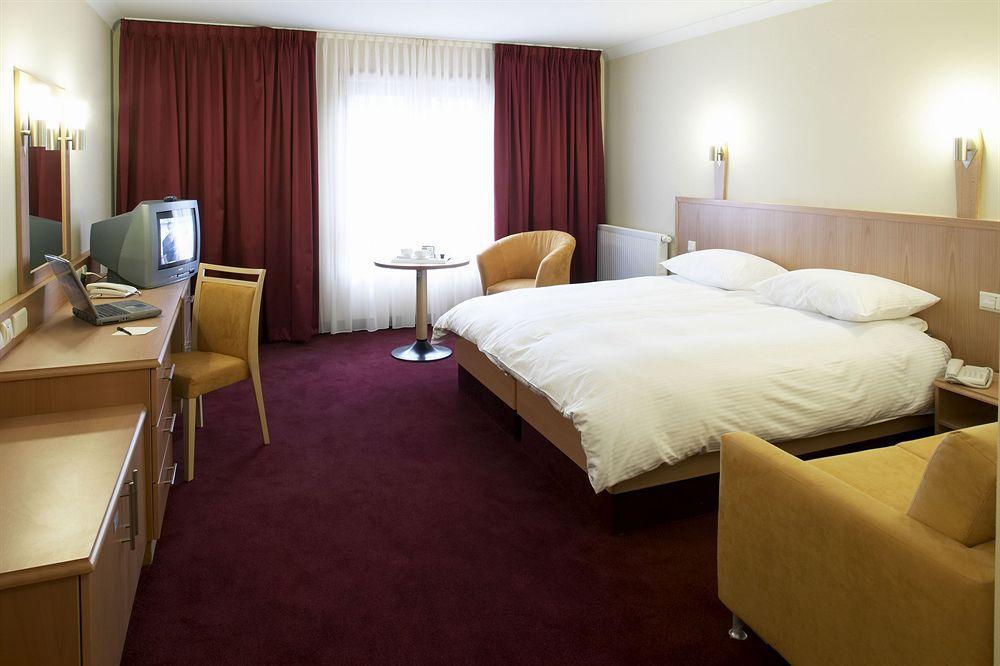 Clayton Hotel Dublin Airport Coolock Oda fotoğraf The photo shows a hotel room featuring a double bed with white linens and pillows. There are two bedside lamps mounted on the wall above the bed. To one side, there's a small round table with two chairs. Opposite the bed, a wooden desk with a chair i